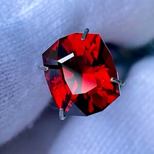 May include: A deep red gemstone with a square cut, held in place by four prongs. The gemstone is set against a white background.