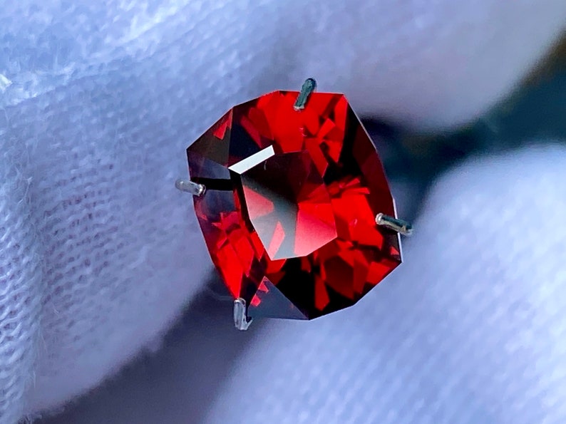 May include: A deep red gemstone with a square cut, held in place by four prongs. The gemstone is set against a white background.