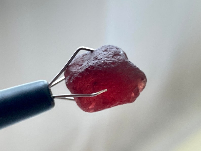 May include: A deep red gemstone held in a metal tweezer. The gemstone is rough and has a natural, unpolished look.