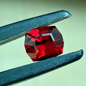 May include: A deep red gemstone with a faceted cut, held by tweezers. The gemstone is a square shape with rounded corners.