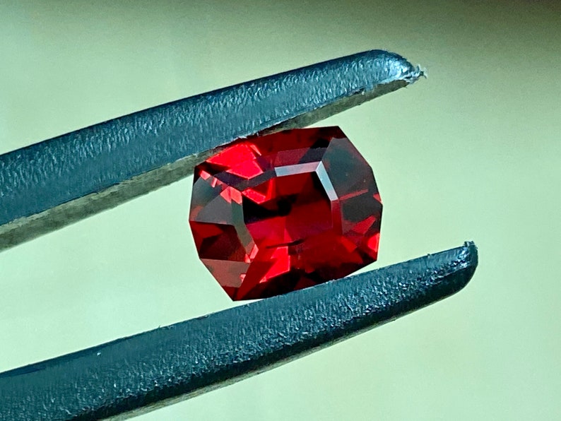 May include: A deep red gemstone with a faceted cut, held by tweezers. The gemstone is a square shape with rounded corners.