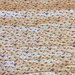 May include: A white fabric with a repeating pattern of small, stylized brown, red, and blue shapes. The fabric has a slightly wrinkled texture.