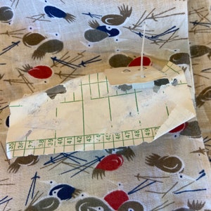 May include: A close-up of a white paper tag with a grid pattern and numbers from 35 to 49. The tag is torn and has a small hole in the center. The tag is attached to a piece of fabric with a white plastic pin.