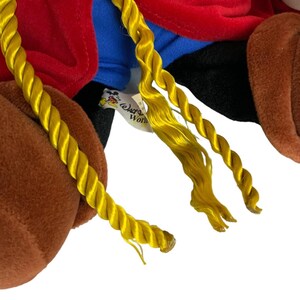 Peut inclure: Un jouet en peluche avec une fourrure brune, des v&ecirc;tements rouges et bleus et de longues tresses jaunes. Le jouet porte une &eacute;tiquette indiquant "Disney World".