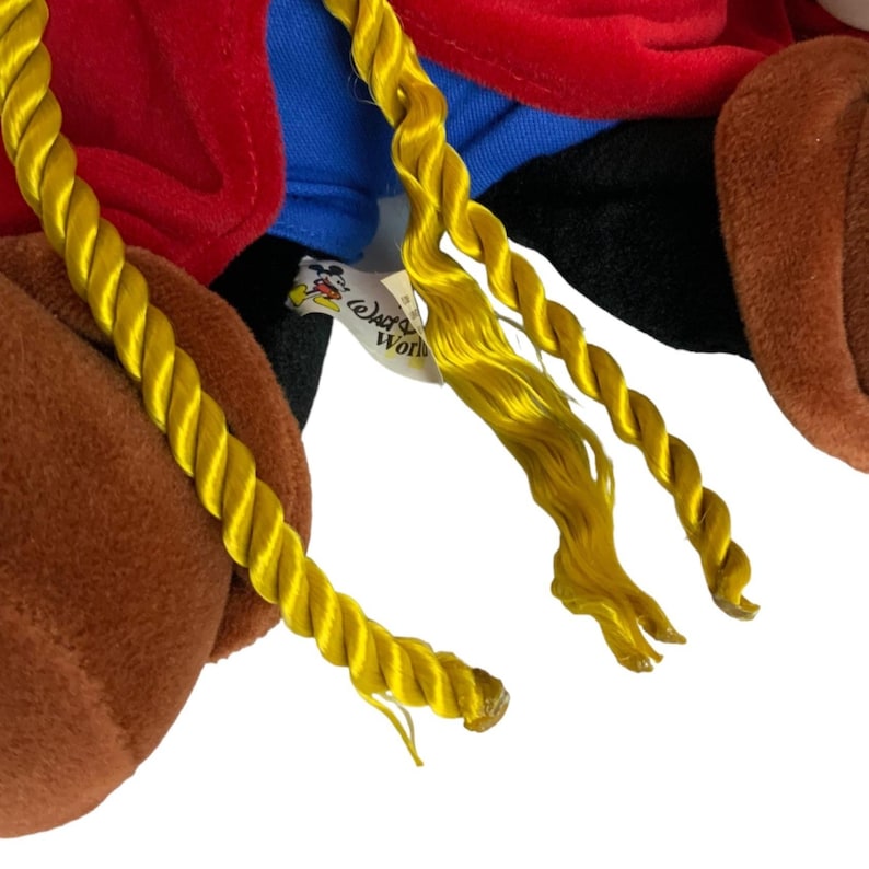 Peut inclure: Un jouet en peluche avec une fourrure brune, des v&ecirc;tements rouges et bleus et de longues tresses jaunes. Le jouet porte une &eacute;tiquette indiquant "Disney World".