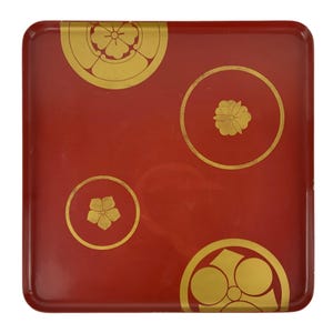 May include: A square red lacquer tray featuring four distinct gold circular motifs. Each gold design depicts a different stylized floral or geometric pattern, including a five-petaled blossom and circular arrangements, set against the smooth red surface.
