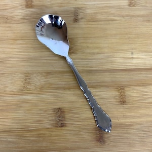 Può includere: Un cucchiaino da zucchero d'argento con un manico decorativo. Il cucchiaino ha una ciotola arrotondata e un bordo smerlato.