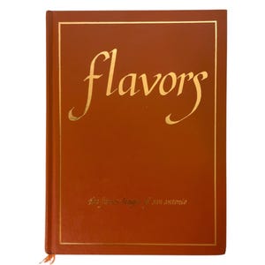 May include: A hardcover cookbook with a brown cover and gold lettering that reads "flavors". The title is in a cursive font. The text "The Junior League of San Antonio" is printed in gold at the bottom of the cover.