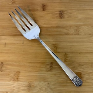 May include: A silver serving fork with four tines. The handle has a floral design at the base. The fork is displayed on a wooden surface.