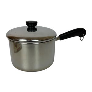 May include: A stainless steel saucepan with a lid and a black handle. The pot has a shiny, reflective surface and a black knob on the lid. The handle is curved and has a small loop at the end.