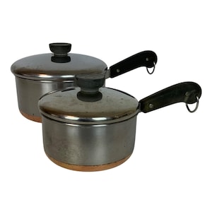 Può includere: Due pentole vintage in acciaio inossidabile con coperchi. Le pentole hanno fondi in rame, manici neri e piccoli anelli di metallo. I coperchi hanno pomelli grigio scuro. Le pentole sono su sfondo bianco.