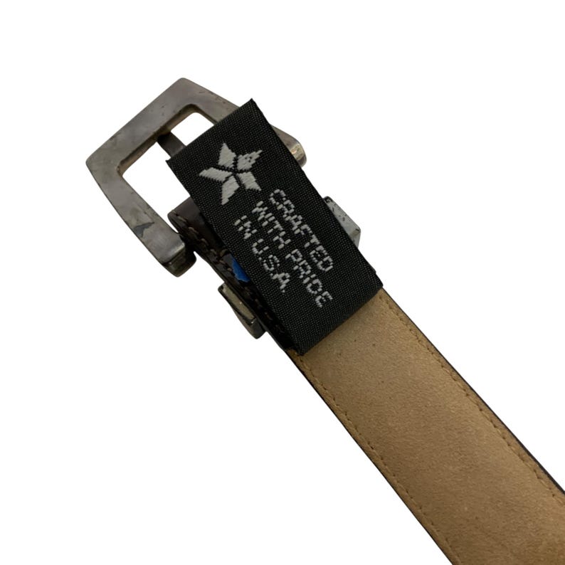 May include: Close-up of a brown leather belt with a silver-colored buckle. The belt has a tag that reads "Crafted with Pride in U.S.A." The belt is likely designed for everyday wear and is a classic accessory.