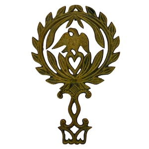 Könnte beinhalten: Ein gusseiserner Topfständer mit goldener Oberfläche. Der Topfständer zeigt einen stilisierten Adler mit einem Herzen in der Brust, umgeben von einem Kranz aus Blättern.