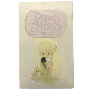 May include: A new, sealed Precious Moments Bible. The cover features a light-colored illustration of a blonde-haired child holding two lambs, with a small dog nearby. The words "Precious Moments" and "Bible" are printed in a soft purple color.