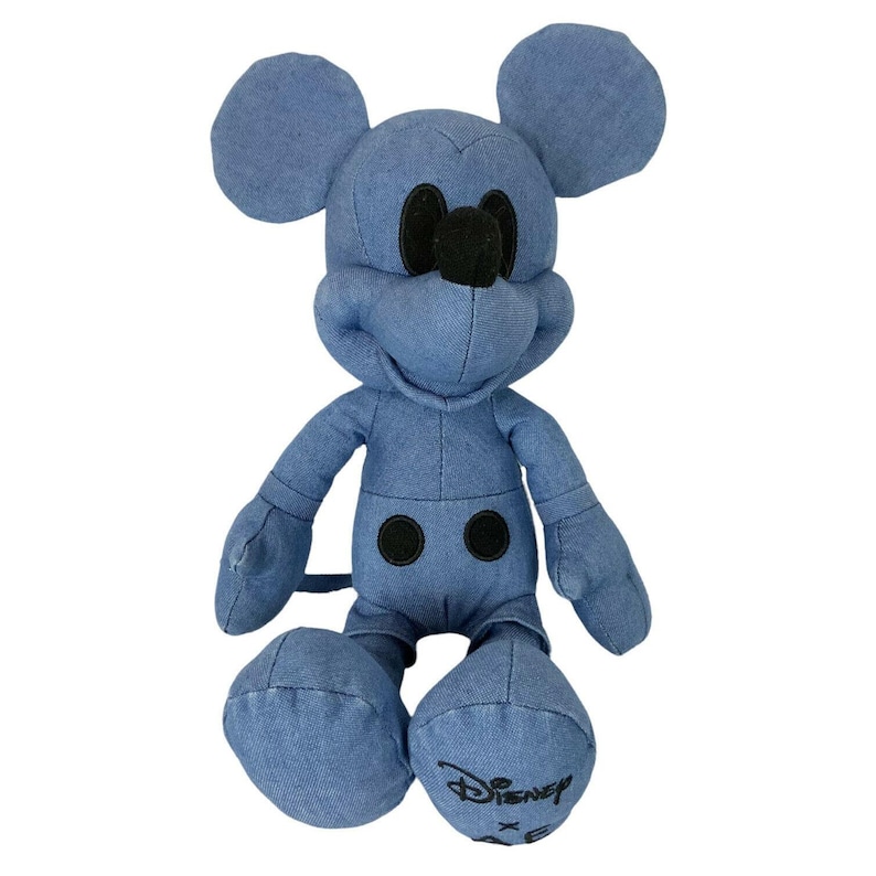 May include: A plush Mickey Mouse character toy constructed from blue denim fabric. The figure features large rounded ears, a black nose, and two black circular buttons on the torso. The left foot displays the black embroidered text Disney x AE.