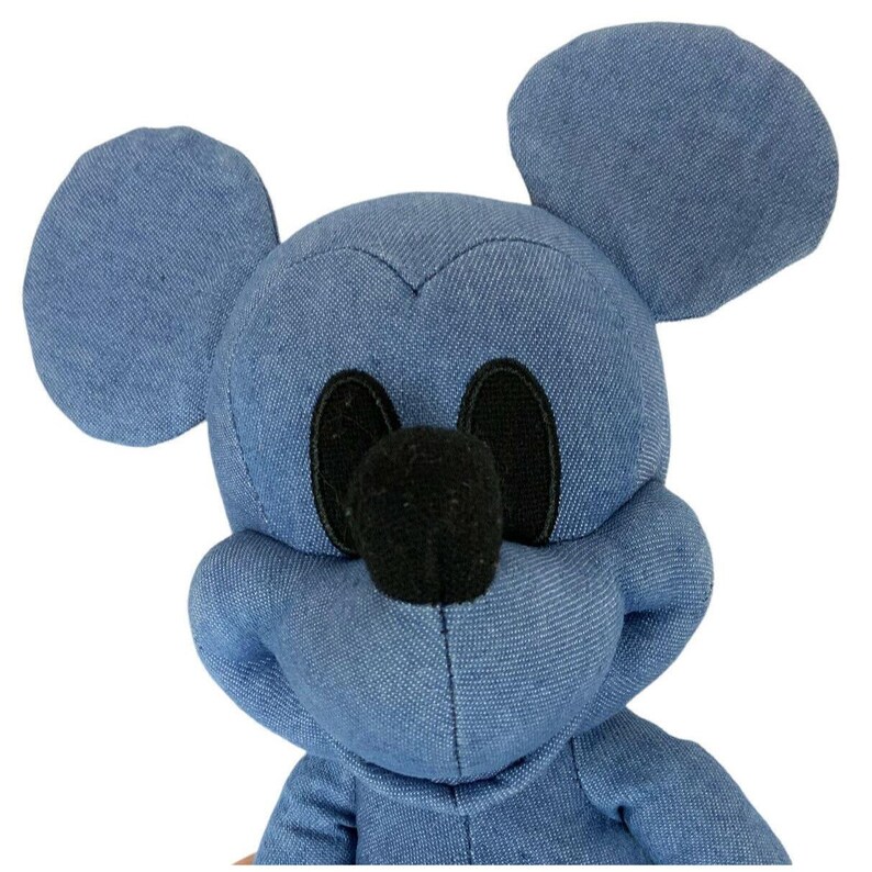 May include: A plush toy shaped like a cartoon mouse head with large rounded ears. The exterior is crafted from a blue denim-like fabric with a visible woven texture. It features two oval black fabric eyes and a prominent rounded black fabric nose.