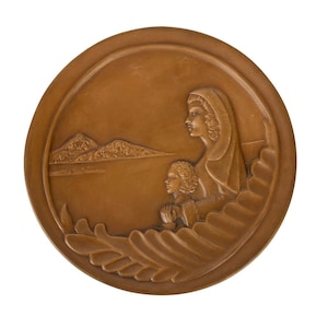 Puede incluir: Una placa circular marrón con un diseño en relieve de una mujer y un niño mirando hacia una cordillera en la distancia. La mujer lleva una túnica larga y el niño tiene las manos juntas en oración.