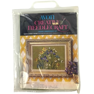 Op de afbeelding: Een Avon Creative Needlecraft crewel borduurpakket genaamd Burst of Spring in een doorzichtige plastic zak. De verpakking toont een afgewerkt bloemmotief in een gouden lijst, met tekst die een stofmaat van 38,1 cm bij 48,26 cm aangeeft.
