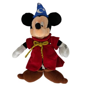 Peut inclure: Un Mickey Mouse en peluche habill&eacute; en sorcier. Il porte une robe rouge avec une ceinture en corde dor&eacute;e, un chapeau bleu avec des &eacute;toiles et une lune croissante, et des chaussures marrons.