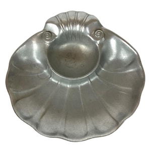 Puede incluir: Un plato de servir de metal plateado con forma de concha, borde acanalado y dos remolinos decorativos en la parte superior.