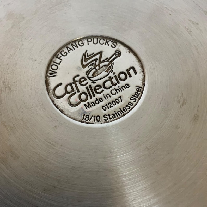Op de afbeelding: Close-up van de bodem van een roestvrijstalen pan met de tekst "Wolfgang Puck's Cafe Collection". De tekst "Made in China" en "18/10 Stainless Steel" zijn ook zichtbaar. De pan heeft een geborstelde metalen afwerking.