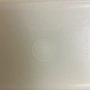Pode incluir: Uma foto em close-up de uma tampa de recipiente de pl&aacute;stico branco. A tampa tem uma superf&iacute;cie ligeiramente texturizada e uma reentr&acirc;ncia circular no centro. A palavra "Tupperware" est&aacute; em relevo na tampa.
