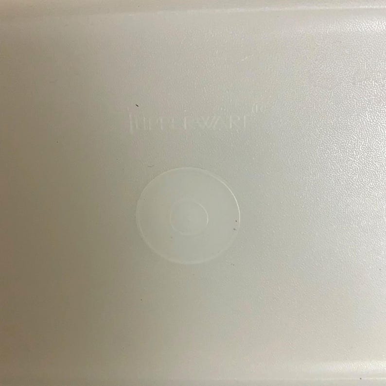 Pode incluir: Uma foto em close-up de uma tampa de recipiente de pl&aacute;stico branco. A tampa tem uma superf&iacute;cie ligeiramente texturizada e uma reentr&acirc;ncia circular no centro. A palavra "Tupperware" est&aacute; em relevo na tampa.