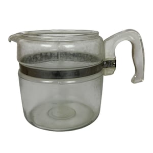 May include: Clear glass coffee pot with a handle and a metal band around the middle. The pot has a spout for pouring and is designed for brewing coffee. The handle is curved and attached to the side of the pot.