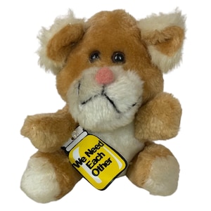 Puede incluir: Un juguete de peluche marrón y blanco con una etiqueta que dice "We Need Each Other". El juguete tiene una nariz rosa y ojos negros.