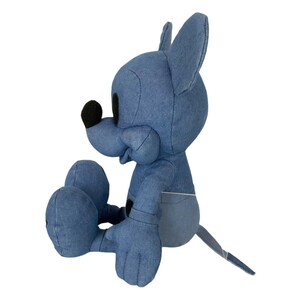 May include: A plush toy character made of blue denim fabric, featuring large pointed ears, a rounded snout with a black nose, and a long thin tail. The figure is seated in a profile position, showcasing stitched seams and a two-tone blue design.
