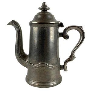 May include: A metallic silver-toned coffee pot with a tall cylindrical body, a curved spout, and a decorative handle. The pot features a removable lid with a finial top and a wavy embossed line detail near the base. The text reads Wilton Columbia PA USA.