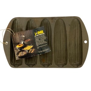May include: A cast iron corn stick pan with five corn-shaped molds. The pan is dark gray and has a tag attached with the Lodge logo and product information. The pan is designed for baking corn sticks.