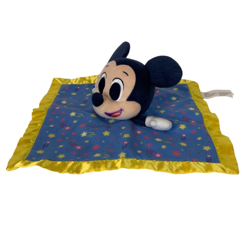 May include: A plush Mickey Mouse security blanket featuring a soft, black and tan character head and one arm attached to a square blue fabric base. The blanket is decorated with a pattern of small yellow stars and pink shapes, finished with a yellow satin border.