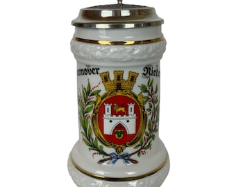 Op de afbeelding: Witte keramische bierkan met een tinnen deksel. De kan heeft een gouden rand en een kleurrijk wapenschild met de tekst "Hannover" en "Niedersachsen".