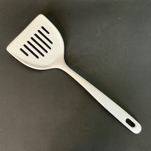 May include: A white slotted spatula with a long handle and a hole at the end for hanging. The spatula head is triangular with several parallel slots. The kitchen utensil is made of a solid, smooth material.