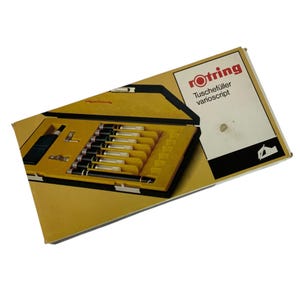 May include: A vintage Rotring Tuschefüller varioscript set in a yellow and black case. The set includes multiple black and silver pens, a bottle of ink, and other accessories. The box has the Rotring logo and product information printed on it.