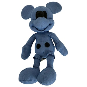 May include: A plush toy shaped like a cartoon mouse with large round ears, a prominent snout, and two black circular buttons on the torso. The entire figure is covered in a solid, muted blue fabric, with a black nose and black circular buttons.