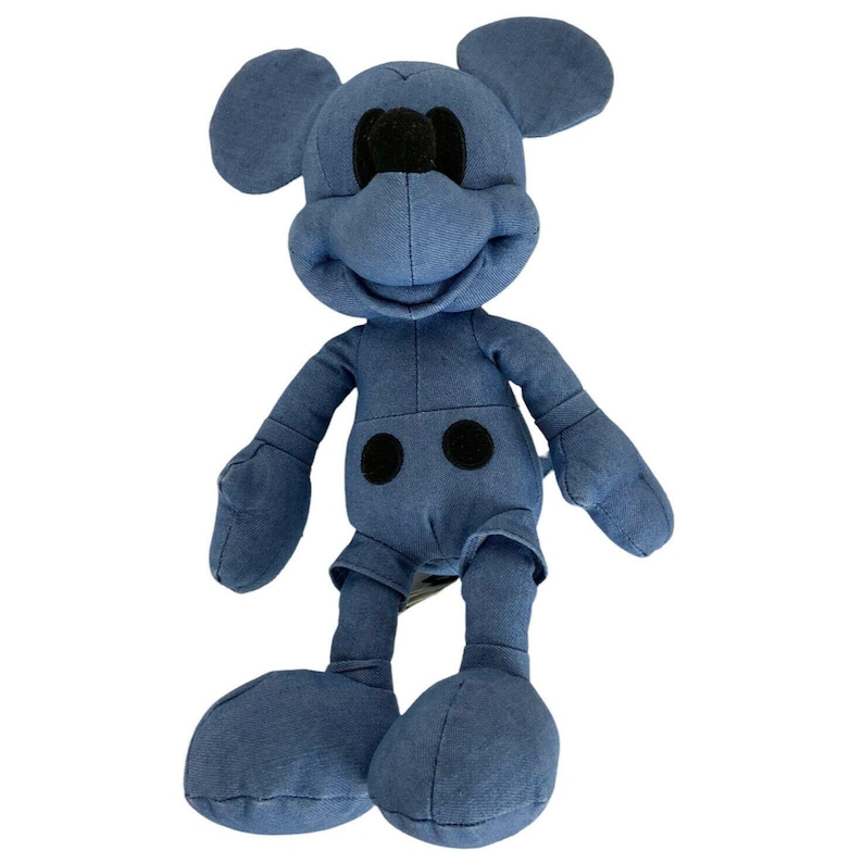 May include: A plush toy shaped like a cartoon mouse with large round ears, a prominent snout, and two black circular buttons on the torso. The entire figure is covered in a solid, muted blue fabric, with a black nose and black circular buttons.