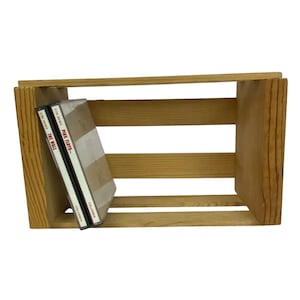 May include: A light brown wooden crate with horizontal slats, holding two CD cases. One CD case is labeled "THE WALL" and the other is labeled "PINK FLOYD". The crate is made of unfinished wood.