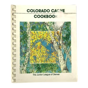 Op de afbeelding: Een spiraalgebonden kookboek met de titel "COLORADO CACHE COOKBOOK" met een kleurrijke omslagillustratie. De omslagkunst toont een schilderij van bomen met gele en groene bladeren tegen een blauwe lucht. De tekst "The Junior League of Denver" staat onderaan.
