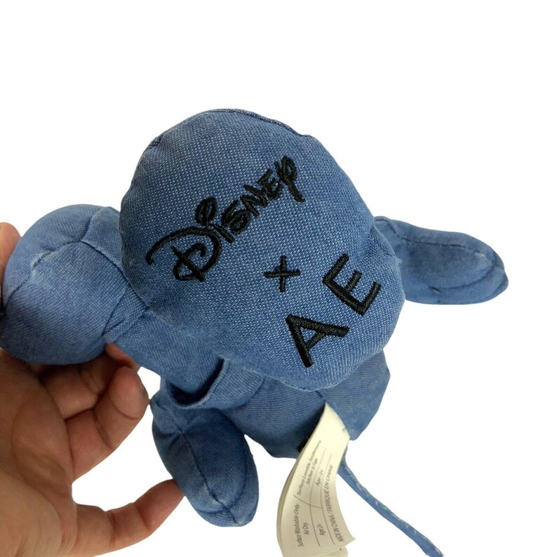 May include: A blue denim plush toy shaped like a mouse with large rounded ears. The back of the head features black embroidered text that reads Disney x AE. A white fabric care tag is attached to the lower portion of the plush toy's body.