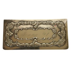 Puede incluir: Una caja rectangular plateada con un diseño grabado ornamentado. La caja tiene una inscripción en relieve que dice "Jenna 91".