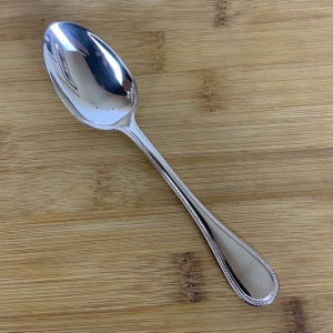 Puede incluir: Una cuchara de plata con un borde de cuentas y un cuenco redondeado. La cuchara está sobre una superficie de madera.