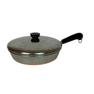 Puede incluir: Una sartén de acero inoxidable con tapa y mango negro. La sartén tiene una base de color cobre y un pomo negro en la tapa. El mango tiene un lazo para colgar.