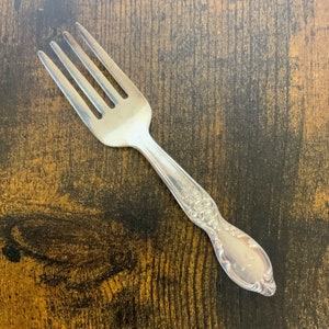 Può includere: Una forchetta d'argento con un designo ornamentale sul manico. La forchetta è su una superficie di legno marrone.