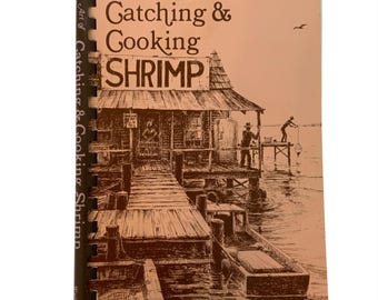 May include: A vintage cookbook titled "The Art of Catching & Cooking Shrimp" with a sepia-toned illustration of a dockside scene. The cover features a wooden pier, a small boat, and figures fishing. The author's name, Lynette L. Walther, is at the bottom.