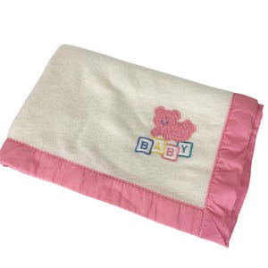 May include: A soft, off-white baby blanket with a pink satin border. The blanket features an embroidered design of a pink teddy bear and the word "BABY" in colorful block letters. The blanket is folded neatly.