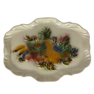 May include: A white ceramic serving tray with a raised design of fruit. The tray features a pineapple, bananas, grapes, apples, and pears in shades of green, yellow, red, and purple.