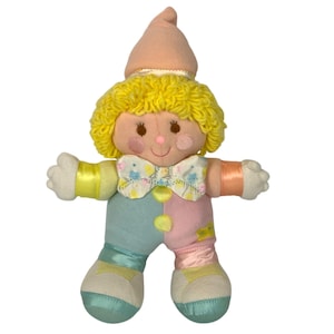 May include: A vintage plush doll in a clown design. The doll has a peach-colored pointed hat, yellow yarn hair, and a bow tie with a floral pattern. The doll's body is divided into sections of blue, pink, and yellow, with matching accents.