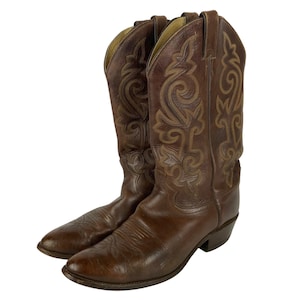 Op de afbeelding: Een paar bruine leren cowboylaarzen met een spitse neus, een hak en ingewikkelde beige stikpatronen langs de hoge schacht. De laarzen hebben treklussen aan de bovenkant en een gladde afwerking op de wreef, wat een klassiek ontwerp toont.